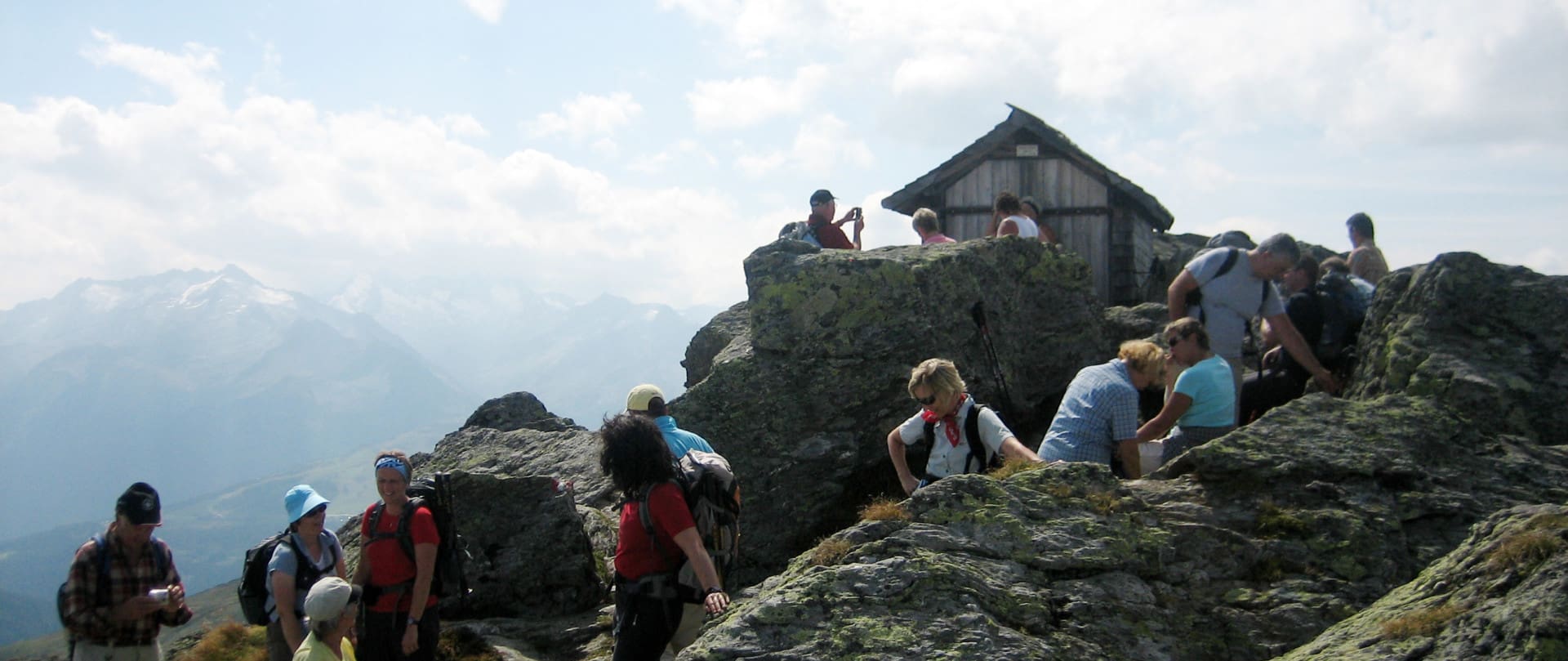 Wanderer erklimmen das steinige Terrain zum Gipfel des Kröndlhorn, wo eine kleine Hütte neben einem Kreuz steht, mit einer beeindruckenden Aussicht auf die verschwommenen Alpen im Hintergrund