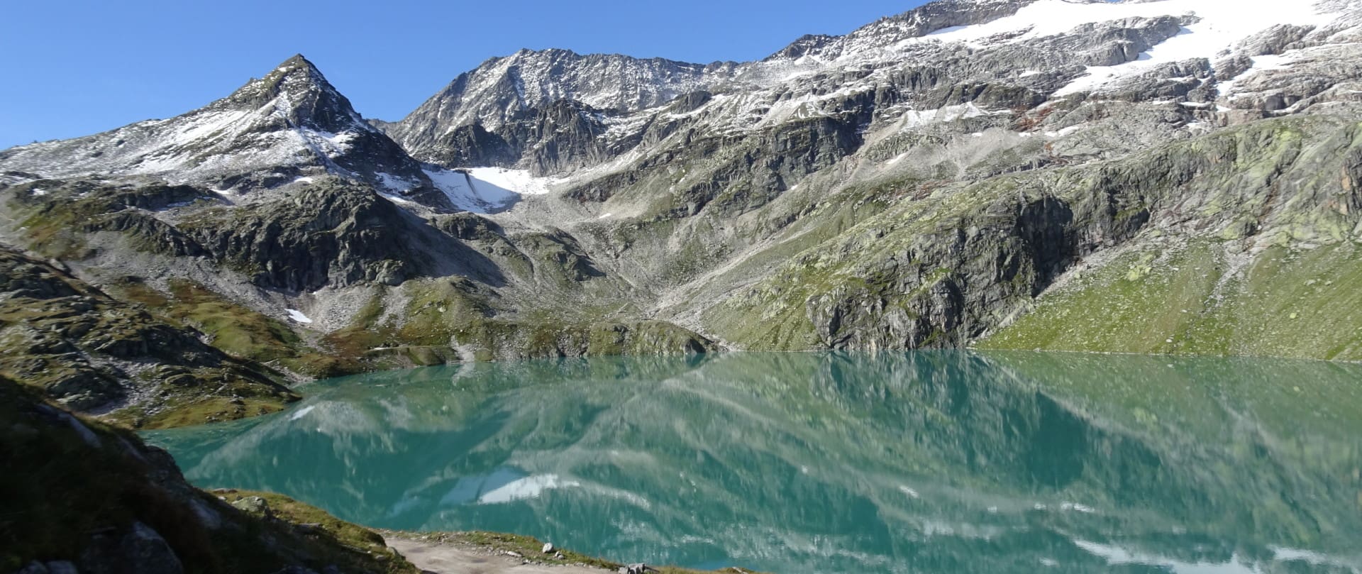 Ein kristallklarer Gebirgssee spiegelt die umliegenden steilen Gipfel und schneebedeckten Hänge unter einem hellblauen Himmel im Weißsee-Gebiet